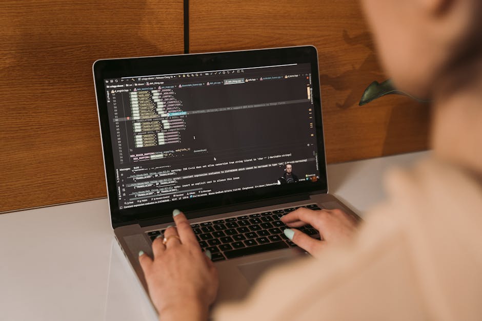A person using a laptop with a coding or programming interface displayed on the screen, sitting at a white desk against a wooden wall background. The laptop screen shows multiple lines of code with sy